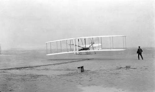 Первый полёт Флайера-1 17 декабря 1903 года, пилотирует Орвилл, Уилбур — на земле. Фотография Джона Т. Дэниелса со спасательной станции Килл Дэвил Хиллс, использован фотоаппарат Орвилла на треножнике